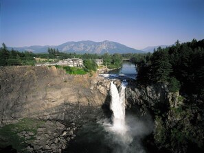 Una cascata che si getta in un fiume, circondata da colline boscose e un edificio con numerose finestre.
