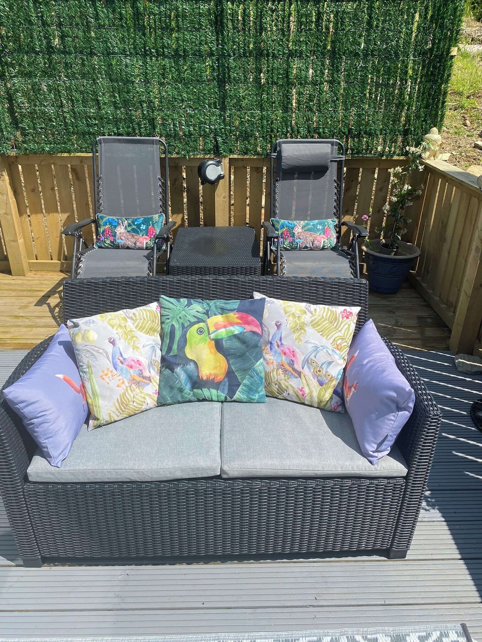 Een rieten bank met kleurrijke kussens met tropische motieven, twee zwarte stoelen met bijpassende kussens, en een kleine tafel op een houten terras.