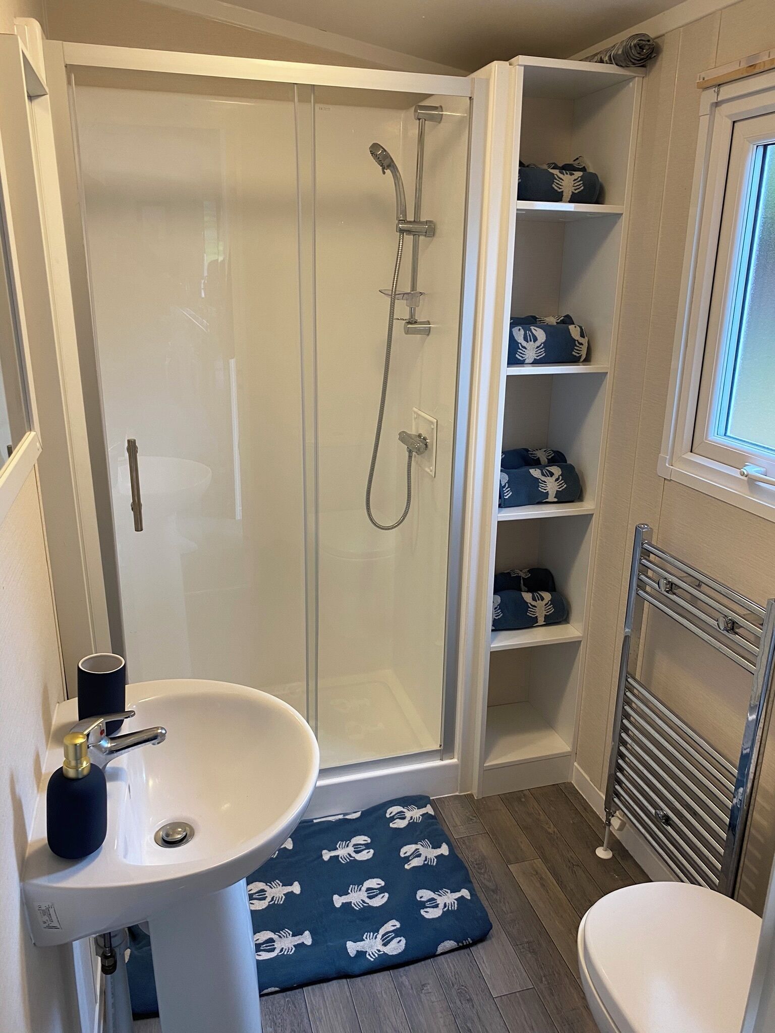Une salle de bain avec une douche à parois de verre, un lavabo, des toilettes et des étagères où sont pliées des serviettes.