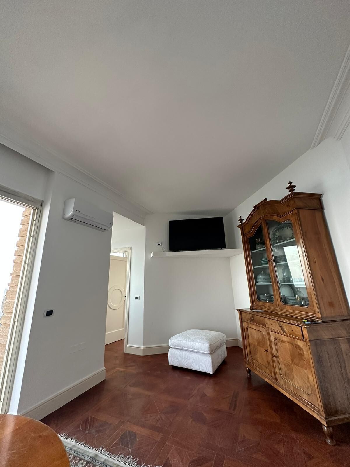 Ein Zimmer mit einem Holzschrank, einem weißen Hocker und einem wandmontierten Flachbildfernseher.