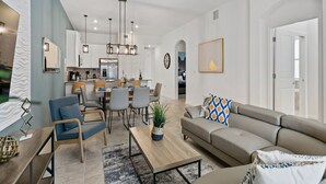 A modern living room with a sofa, coffee table, and chairs. There is a dining area with a table and chairs. The room has a kitchen in the background.