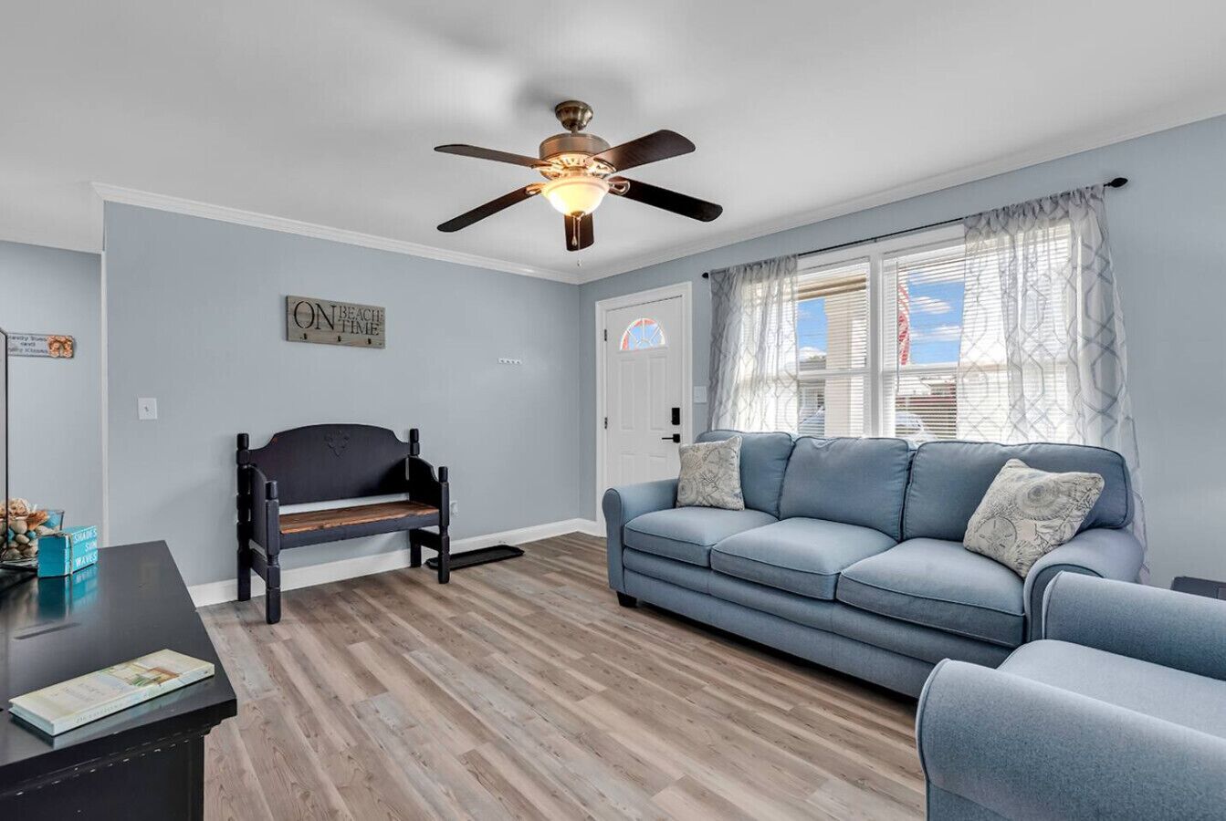 A living room with a blue sofa, a wooden floor, a ceiling fan, and a window with curtains.
