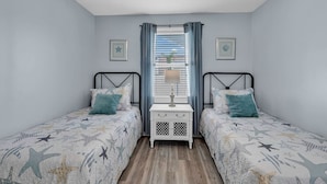 Un dormitorio con dos camas, una mesita de noche, una ventana con cortinas y un ventilador de techo.