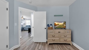 Un pasillo con suelo de madera, pared azul claro y puerta blanca. Se ve un aparador con cajones y un televisor encima. Un cartel en la pared dice "SEASIDE".
