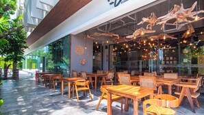 An outdoor seating area with wooden tables and chairs, a modern building facade, and hanging light fixtures. - Ipana by Casago (Playa del Carmen)