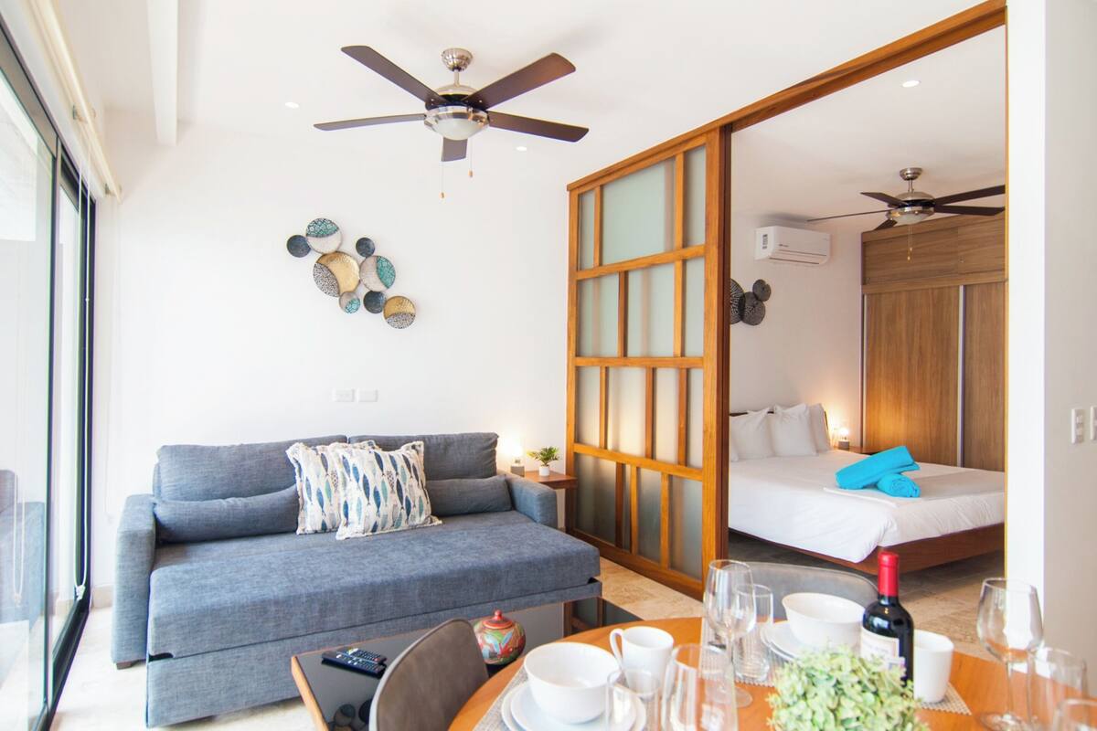 Quarto de hotel moderno com sofá, mesa de jantar posta com copos e garrafa, ventilador de teto e porta deslizante que dá para um quarto.