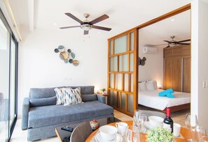 A modern hotel room with a sofa, a dining table set with glasses and a bottle, a ceiling fan, and a sliding door leading to a bedroom. - Ipana by Casago (Playa del Carmen)