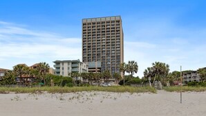 A beachfront hotel with a tall building, a sandy beach, and palm trees.