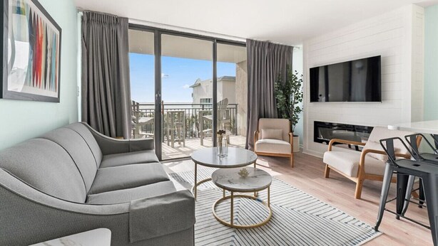 A modern living room with a grey sofa, a round coffee table, a beige armchair, a TV mounted on the wall, and a balcony with a view of buildings.