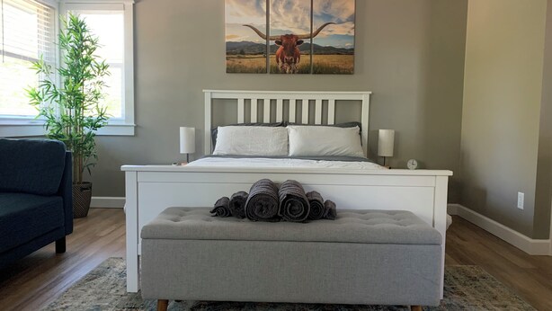 A bedroom with a bed, bedside lamps, a bench with rolled towels, a potted plant, and a large landscape painting on the wall. - The Hyde Park Hideaway | Centrally-Located | Cozy (Austin)