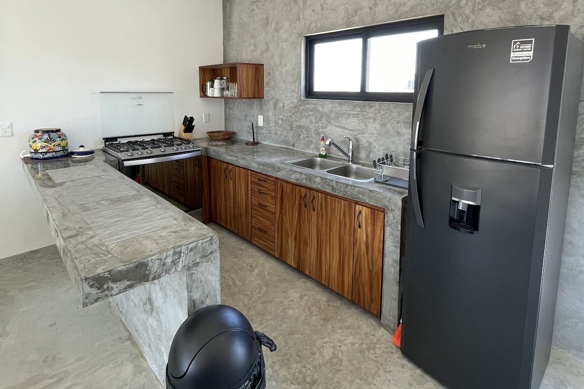 A modern kitchen with a marble countertop, wooden cabinets, a black refrigerator, and a window above the sink.