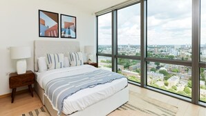 Une chambre à coucher avec un grand lit, une table de chevet, une lampe et une vue sur le paysage urbain, visible à travers de grandes fenêtres.
