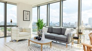 Un salon moderne avec un canapé blanc et gris, un fauteuil blanc et une table basse. Une grande fenêtre offre une vue sur la ville.
