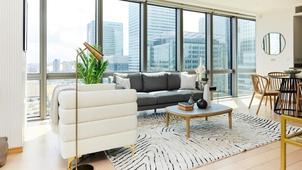 Un salon moderne avec un canapé, une table basse et des fauteuils, de grandes fenêtres offrant une vue sur la ville, et une plante en pot.