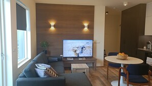 Una sala moderna con un televisor de pantalla plana grande, un sofá gris, una mesita de centro de madera y un comedor con una mesa redonda y sillas.