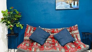 Camera da letto moderna con una parete blu a contrasto, un dipinto con una veduta cittadina, un divano grigio con cuscini decorati e una pianta in vaso.