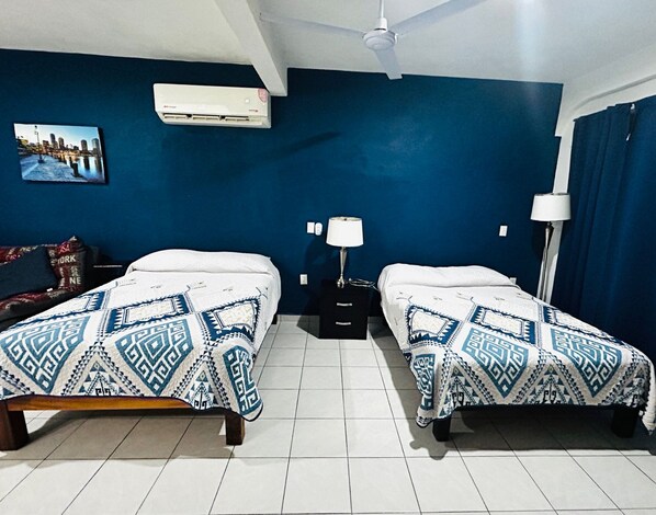 A hotel room with two beds, a ceiling fan, an air conditioner, a nightstand with a lamp, and a wall-mounted picture.