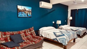 Camera d'albergo con una parete blu, un quadro che raffigura un panorama urbano, un divano con cuscini decorati, due letti con coperte a motivi e una ventola a soffitto.