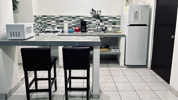 Una cucina compatta con forno a microonde, tostapane, bollitore e un piccolo frigorifero. Ci sono due sedie nere e un lavandino con rubinetto.