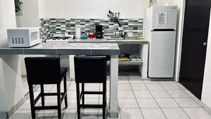 Cocina compacta con microondas, tostadora, hervidor de agua y un refrigerador pequeño. Hay dos sillas negras y un lavamanos con grifo.