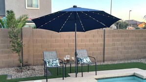 A swimming pool with two lounge chairs and a table, a blue umbrella, and a brick wall in the background. - Aris Oasis  Private Pool Heat Included (Gilbert)