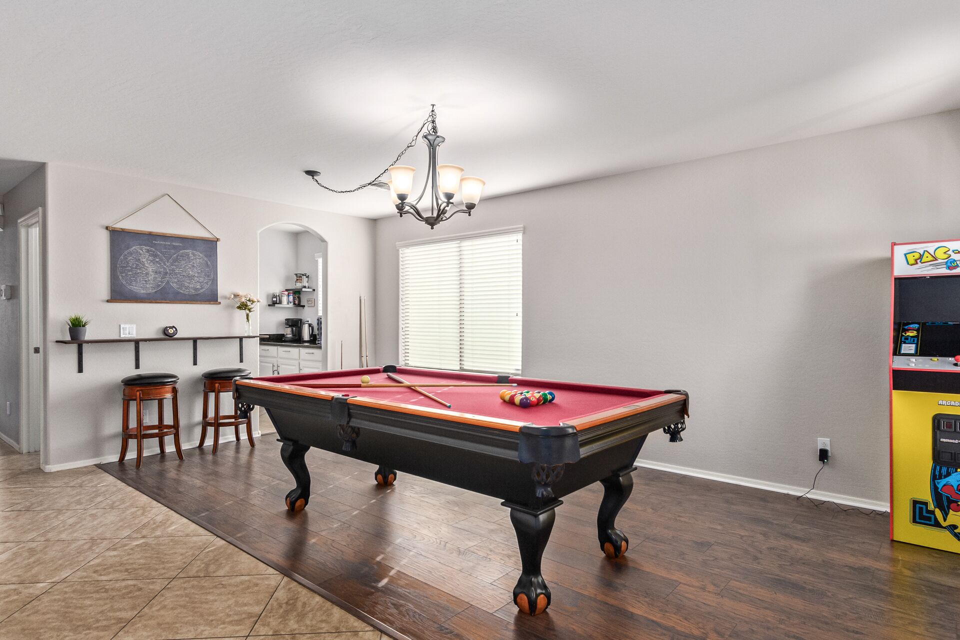 A pool table in a room with wooden flooring, a chandelier, a bar area with stools, and a pinball machine.