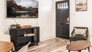 Una sala de estar con una televisión montada en la pared, una puerta oscura, un sillón verde, una mesa de centro de madera y un sofá blanco.