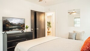 Un dormitorio con una cama, un televisor sobre un tocador, una puerta y un ventilador de techo.