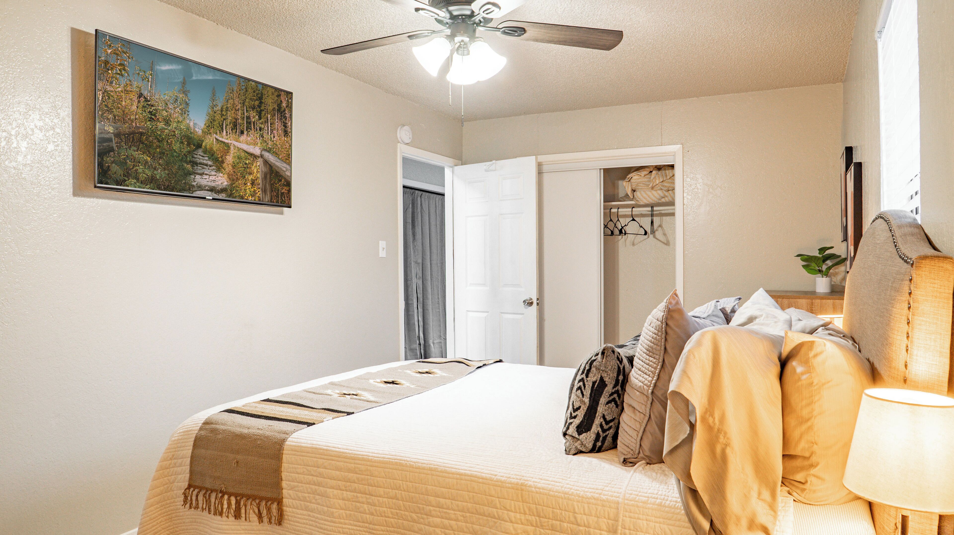 A bedroom with a bed, a ceiling fan, a painting of a forest, and a door leading to a closet.