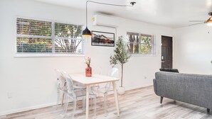 Ein modernes Wohnzimmer mit Essbereich, mit einem Holztisch, klaren Stühlen, einem grauen Sofa und einer Topfpflanze.