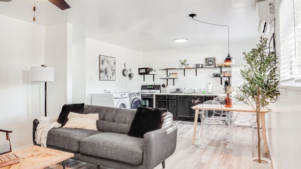 Ein modernes Wohnzimmer mit einem grauen Sofa, einem Couchtisch und einem Essbereich mit klarem Tisch und Stühlen. Im Hintergrund ist eine Küche mit Geräten und einer hängenden Pflanze zu sehen.