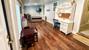 Une pièce compacte et bien éclairée, dotée d’un plancher en bois, d’un bureau avec une chaise, d’un canapé, d’un téléviseur fixé au mur et d’une petite table à manger avec des chaises.