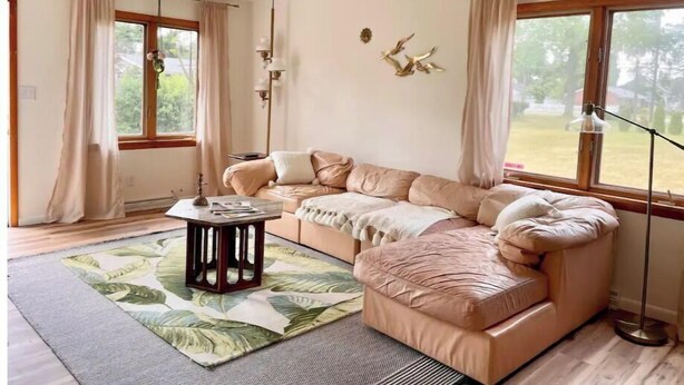 A cozy living room with a sectional sofa, a coffee table, and a floor lamp. There are large windows with curtains, a decorative wall piece, and a view of the outdoors. - The Gemini (Elkhart)