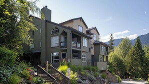 Un immeuble à plusieurs étages, doté d’une façade en pierre, d’un escalier menant à l’entrée et offrant une vue sur les montagnes en arrière-plan.