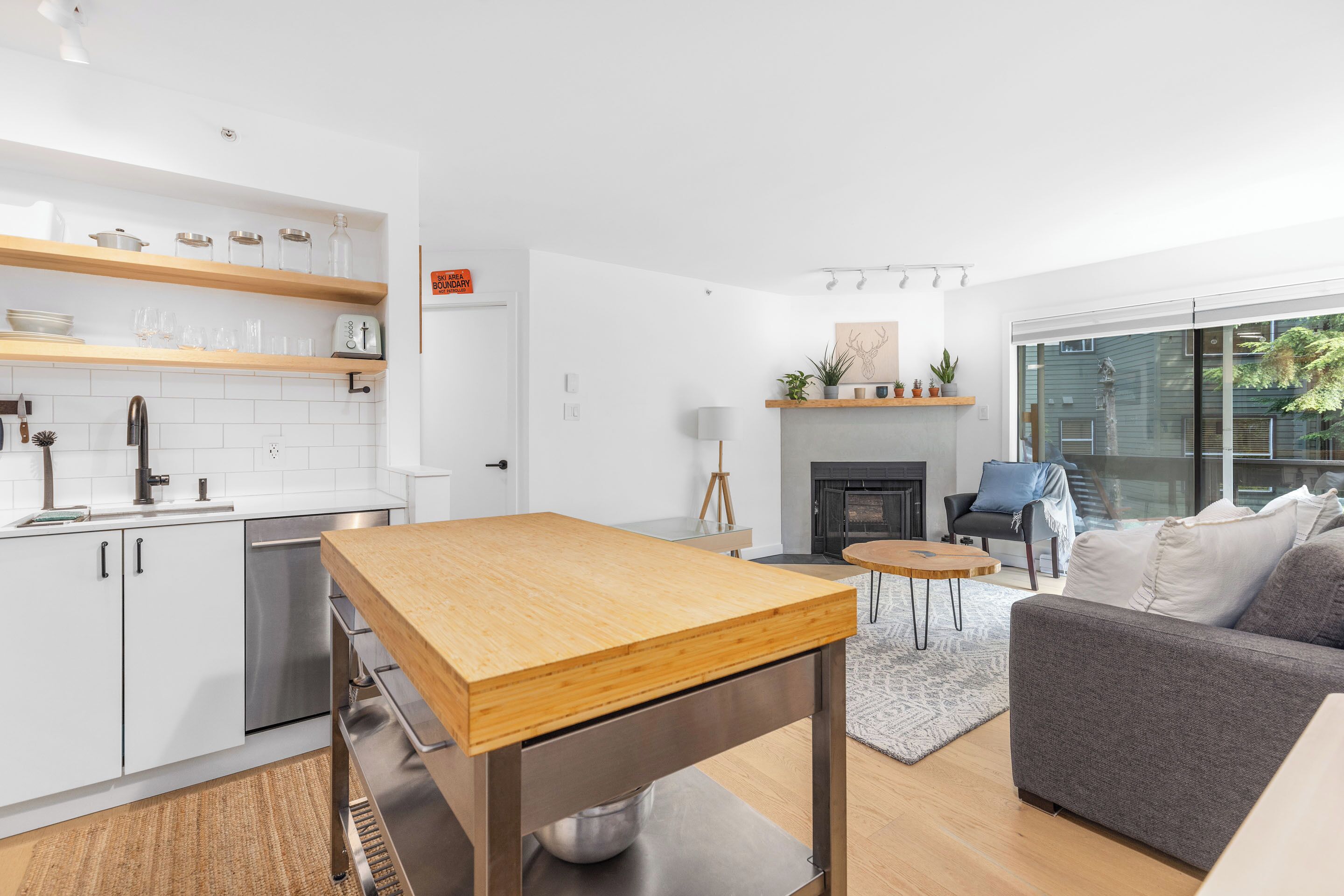 Een moderne keuken met witte kasten, een roestvrijstalen vaatwasser en een houten tafel. In de woonruimte staat een grijze bank en een kleine ronde tafel.