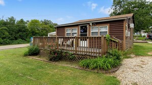 A small, single-story log cabin with a porch, surrounded by a wooden fence and a gravel driveway.