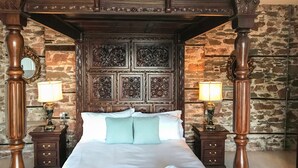 A four-poster bed with intricate carvings, two bedside tables with lamps, and a stone wall backdrop. - Baronet's Quarters (Lynton)