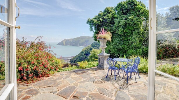 A stone patio with a table and chairs, a vase with plants, and a view of a coastal landscape. - Baronet's Quarters (Lynton)