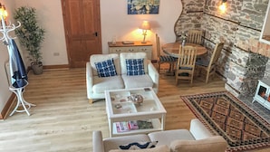 A cozy living room with a sofa, armchair, and coffee table, a dining area with a table and chairs, a stone wall, and a wooden door. - Baronet's Quarters (Lynton)