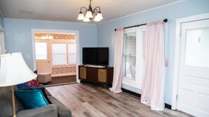 Un salon avec un canapé gris, un téléviseur posé sur un meuble en bois, un lustre et une porte avec une fenêtre.