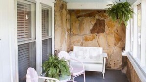 Un porche abrité avec deux chaises blanches et une table, un mur en pierre et une plante suspendue.