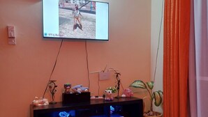 Ein Wohnzimmer mit an der Wand montiertem Fernseher, einer Holztruhe mit Dekoration, einem kleinen Tisch mit Unterlage und einem Blumentopf.