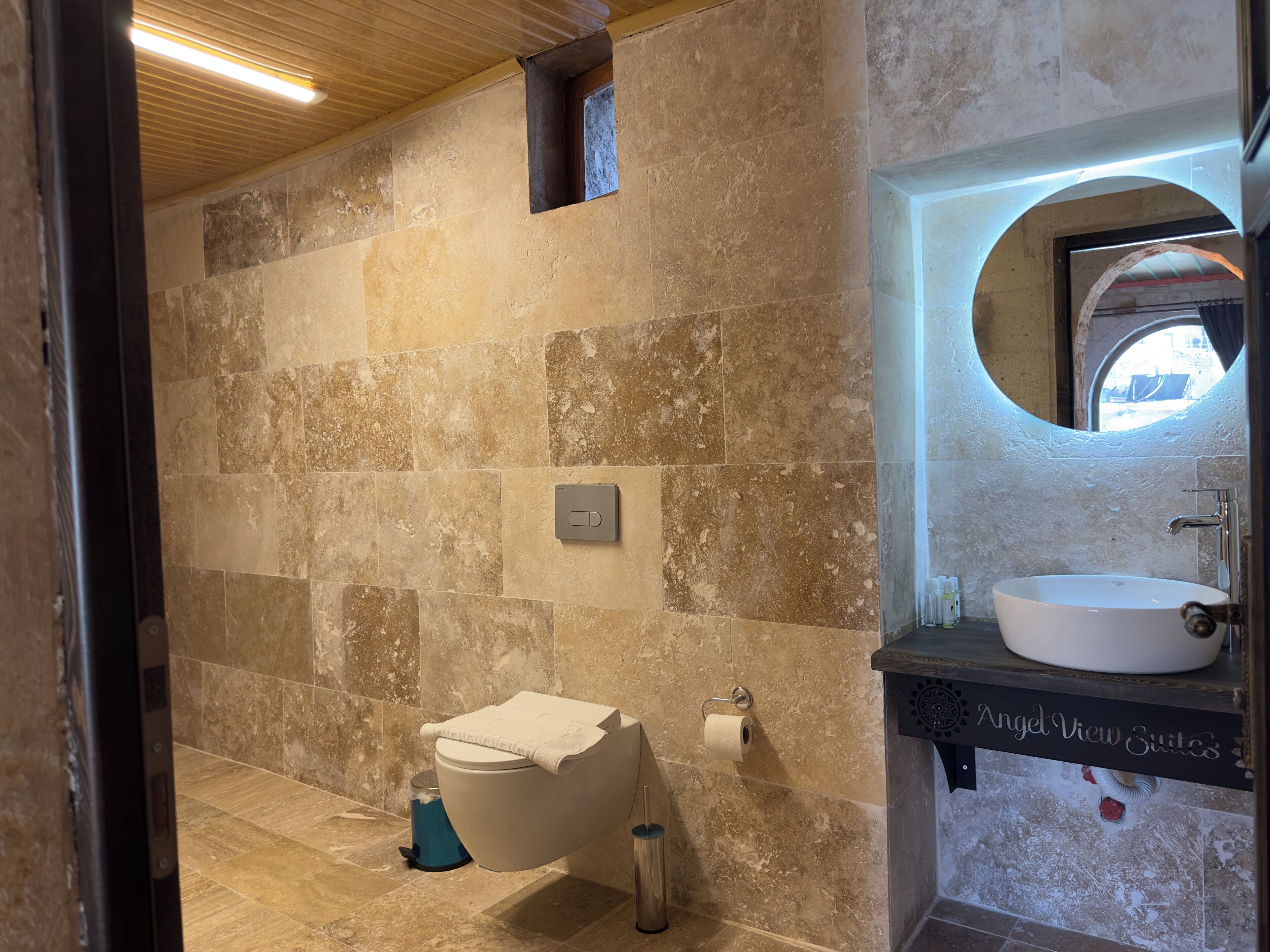 A bathroom with a stone wall, a toilet, a sink with a mirror, and a window.