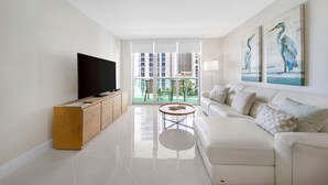 A modern living room with a flat-screen TV, a wooden cabinet, a white sofa, a round coffee table, and a balcony with a view of high-rise buildings. - Luxe Oceanfront Condo in Hollywood Beach (Hollywood)