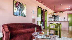 A living room with a red velvet sofa, a glass coffee table, and a green chair. There is a painting on the wall and a kitchen area in the background. - The Leo | In the Heart of Park Circle Main Strip (North Charleston)
