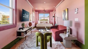 A pink-walled room with a bed, a red sofa, a green chair, a white coffee table, and a TV mounted on the wall. - The Leo | In the Heart of Park Circle Main Strip (North Charleston)