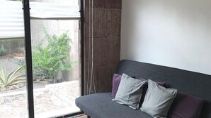 Un salón moderno con un sofá gris, una mesa de madera y un ventanal corredero que da a un área exterior con plantas.