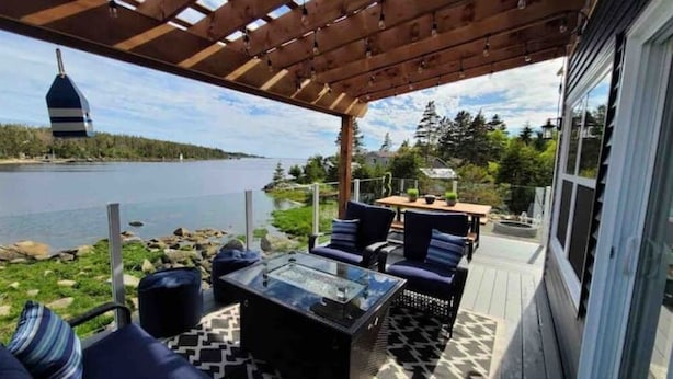 Un patio exterior con una mesa de vidrio y sillas azules, con vista a un lago donde se distingue un faro a lo lejos.