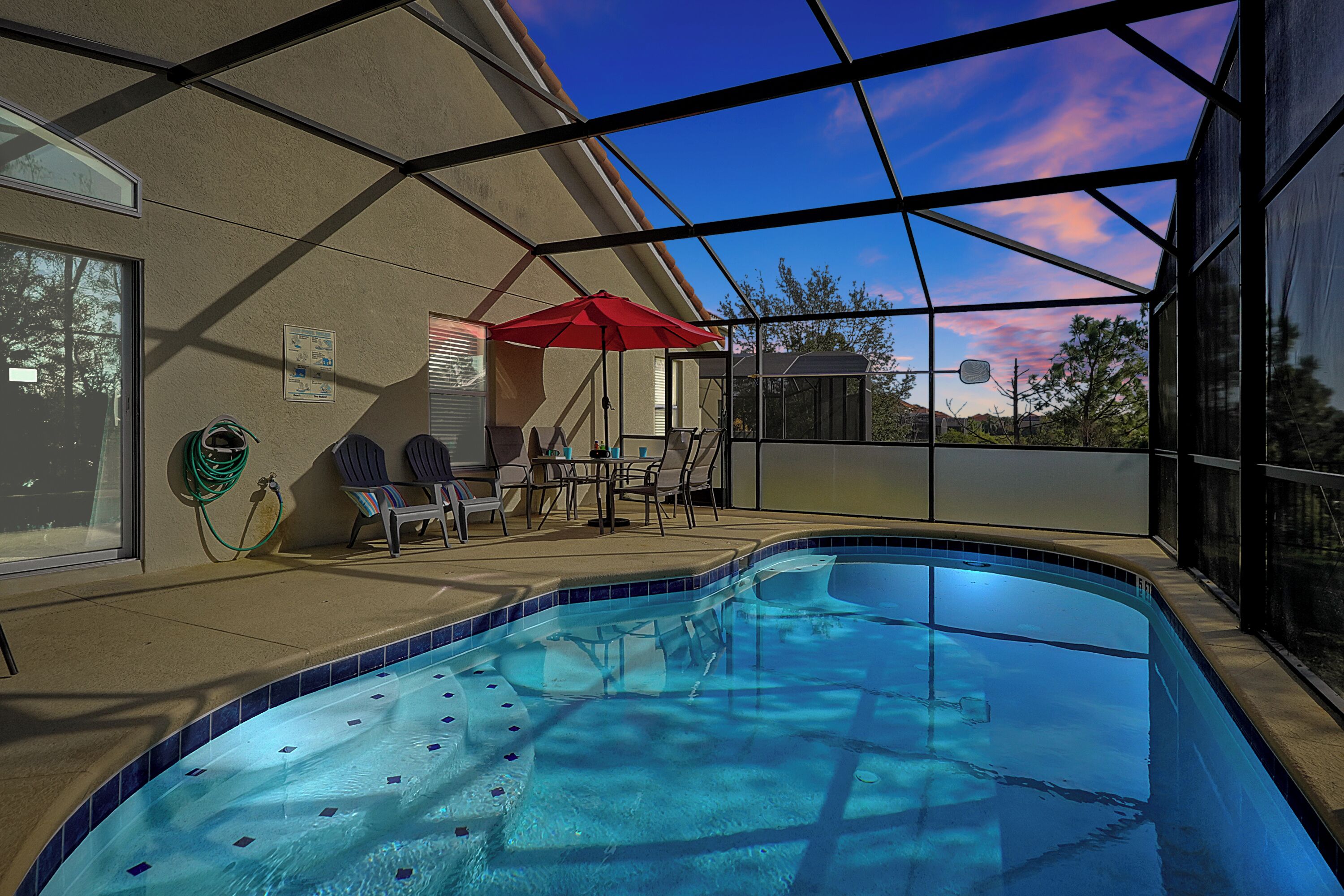 Une piscine entourée de parois en verre, un parasol rouge et des sièges d’extérieur.