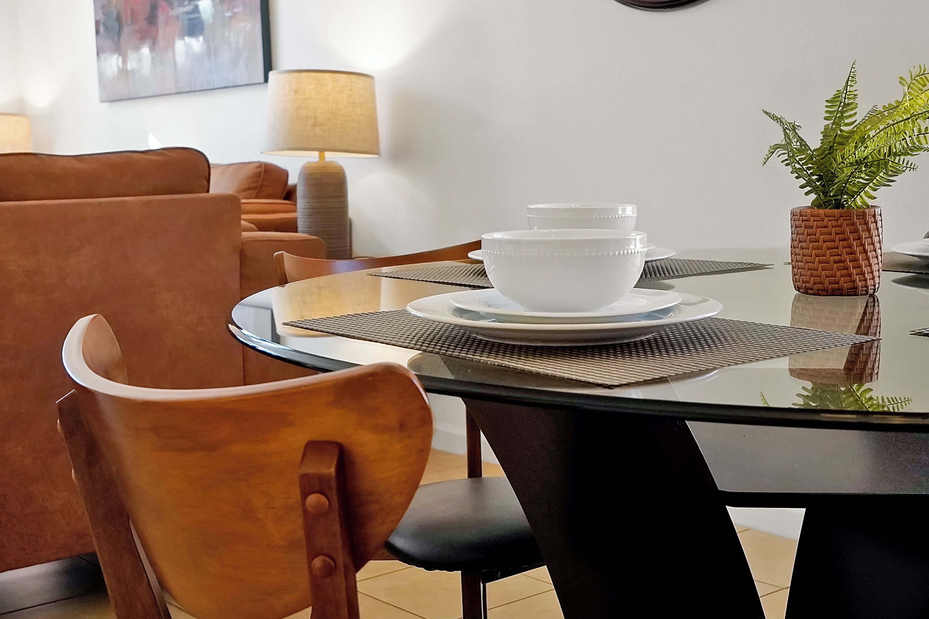 A dining area with a glass table, two white bowls, a woven plant pot, and a brown leather chair.